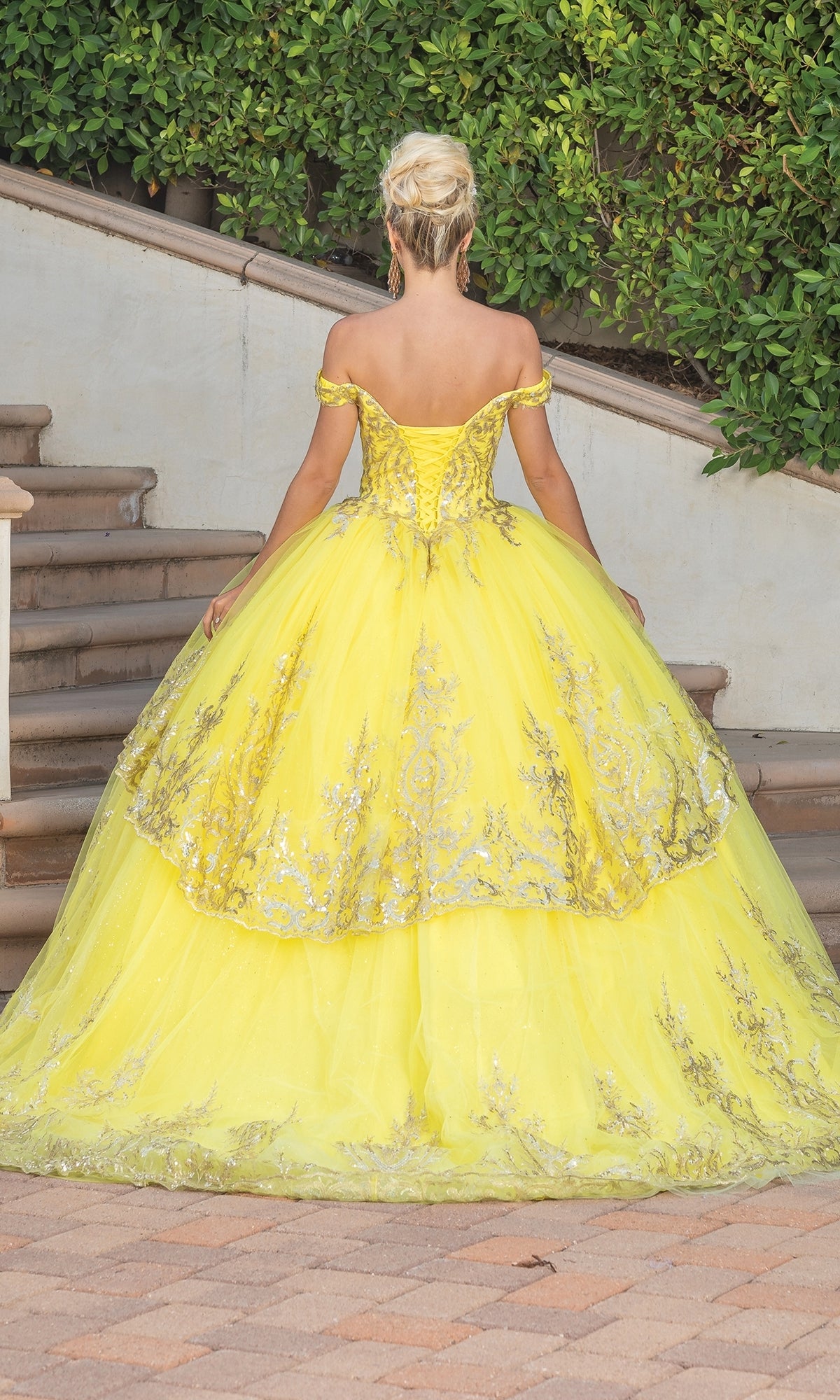 A woman with blonde hair in an updo wears the Quinceanera Dress 1729 by Dancing Queen, a yellow ball gown with gold lace embroidery, standing outdoors near steps and greenery, facing away from the camera.